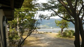 A view of a lake with mountains in the background, a paved area in the foreground, and trees. - Youth Hostel WANIHIMA~Lakeside BIWA (Otsu)
