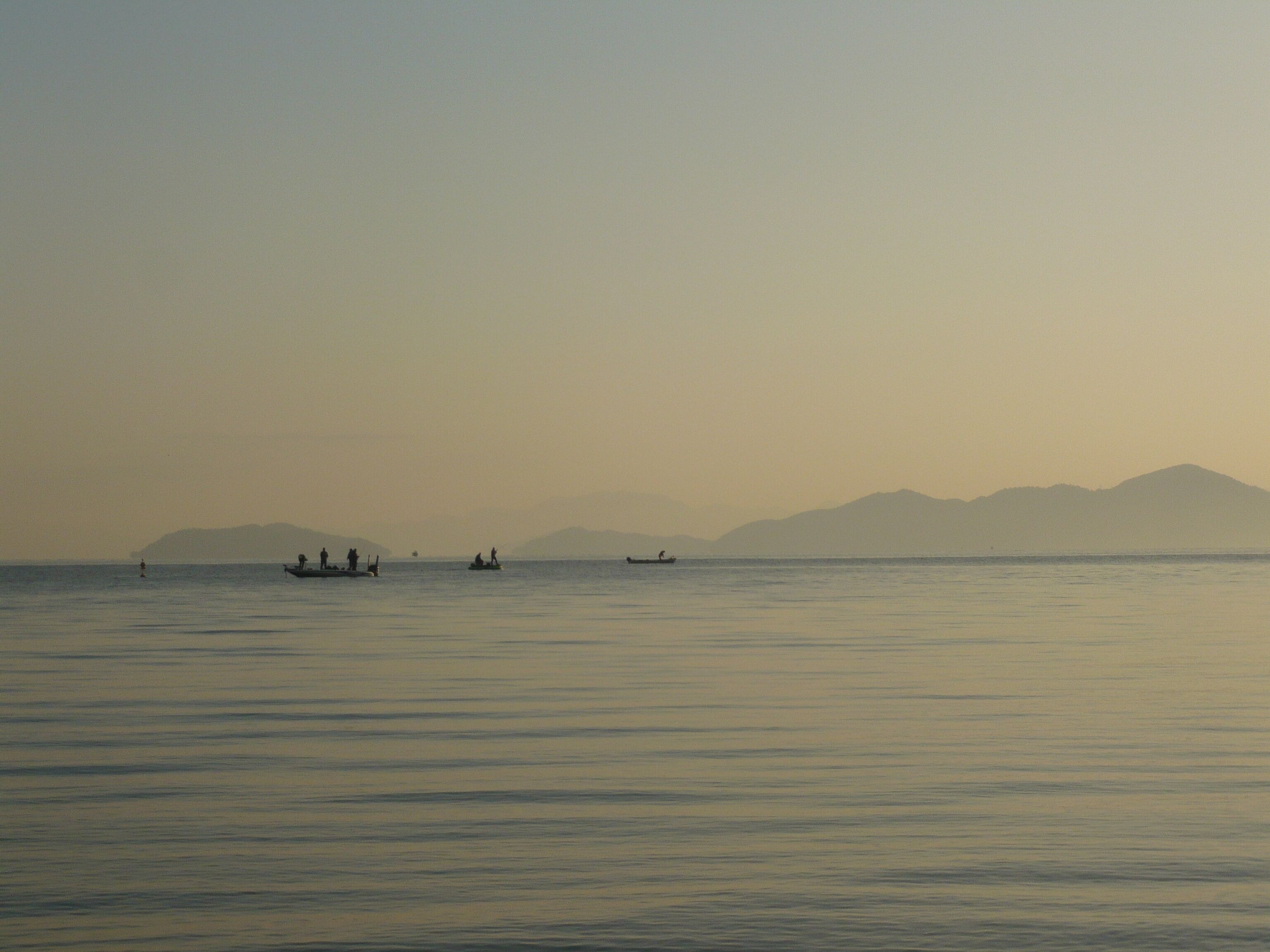 A calm body of water with several boats and distant mountains under a hazy sky.