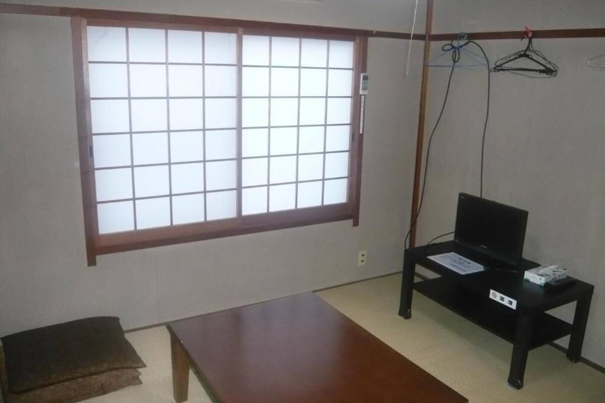 Une pièce dotée d’un sol recouvert de tatamis, d’une petite table en bois, d’un bureau avec un ordinateur et d’une grande fenêtre à panneaux coulissants de style shoji.