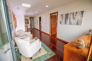 Un couloir avec un fauteuil en cuir blanc, une table en verre ornée d’objets décoratifs, une table d’appoint en bois sur laquelle est posé un objet décoratif, et un tapis à motifs verts.