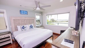A bedroom with a bed, a nightstand, a desk with a chair, a ceiling fan, and a window with a view of the beach.