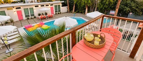 A poolside area with a wooden deck, a red chair, a wicker basket with drinks, and a white umbrella.