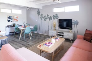 A modern living room with a sofa, a coffee table, a dining area with chairs, a TV, and a wall with a tree mural. - The Rosalie by AvantStay (Palm Springs)