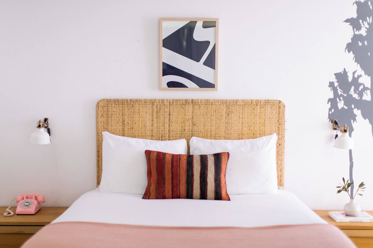 A neatly made bed with white linens and a colorful striped pillow, a woven headboard, a framed abstract artwork above the bed, and a small bedside table with a lamp and a pink telephone.