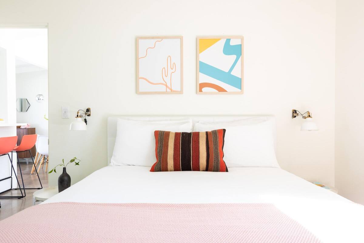 A modern hotel room with a large bed, a desk, and a chair. There are two wall art pieces above the bed.