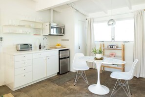 A modern kitchen with white cabinetry, a built-in microwave, and a compact refrigerator. There is a dining area with a white table and two chairs, and a window with curtains. - The Rosalie by AvantStay (Palm Springs)