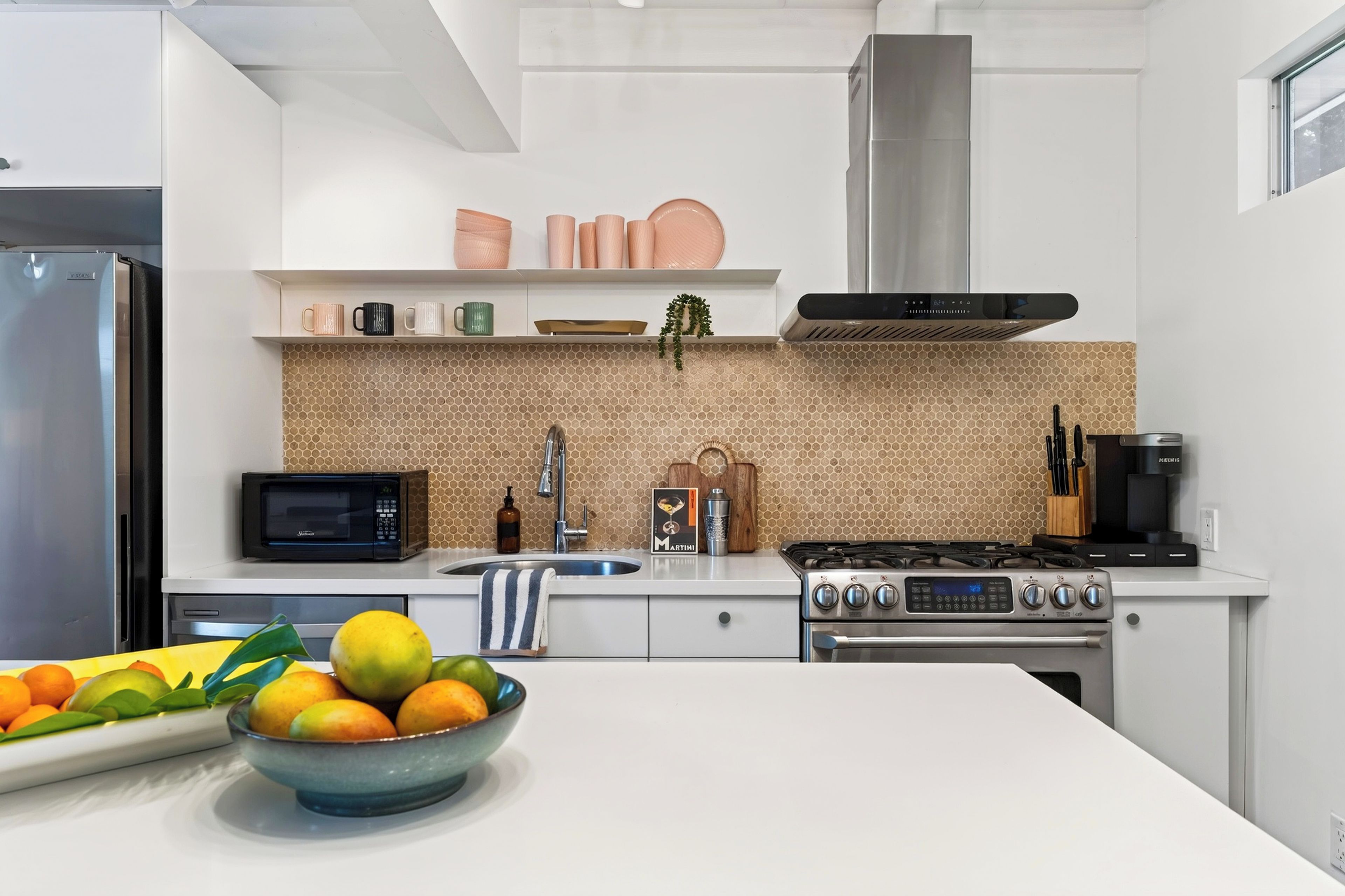 A modern kitchen with a stainless steel refrigerator, a microwave, a gas stove, and a sink. There is a bowl of fruit on the counter.
