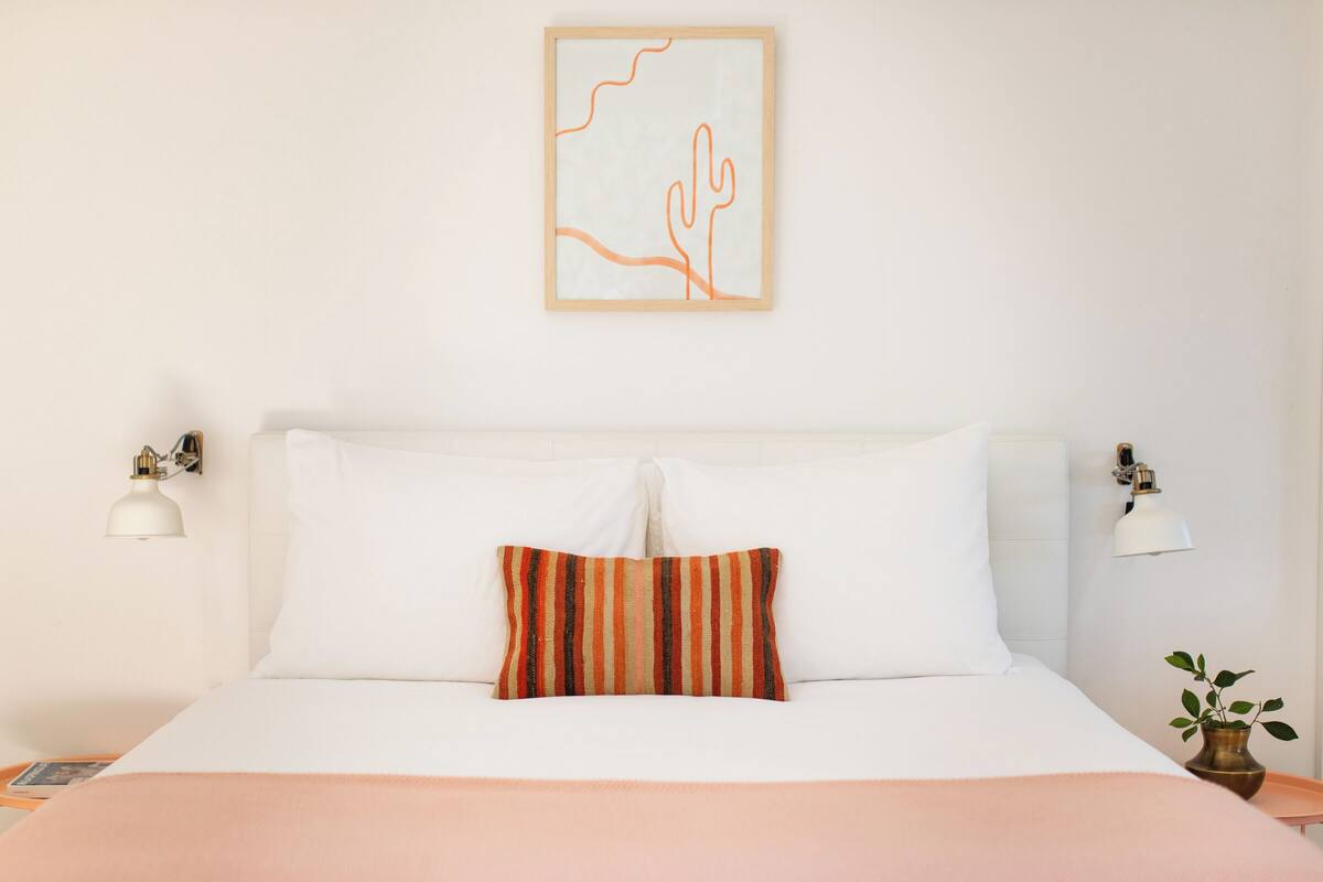 A neatly made bed with white linens and a striped pillow, a framed artwork above it, and bedside lamps on either side.