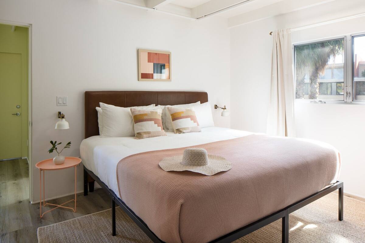A bedroom with a bed, a bedside table with a lamp, a framed picture on the wall, and a window with curtains.