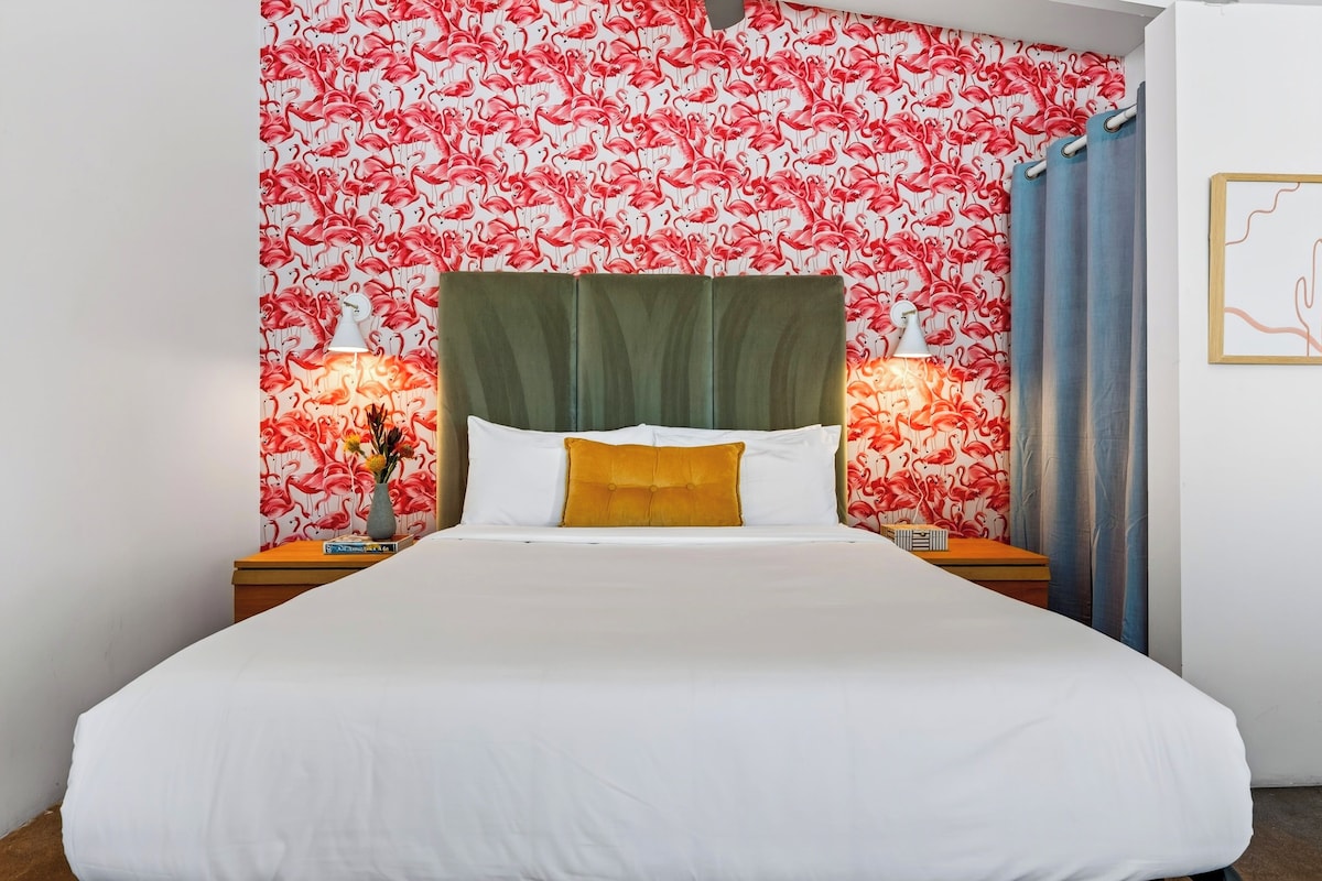 A hotel room with a bed, two bedside tables, a green upholstered headboard, and a wall with a red and white flamingo pattern.
