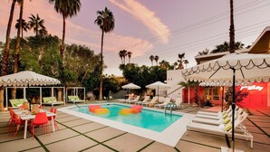 A pool area with lounge chairs and umbrellas, surrounded by palm trees and a building with a red facade. - The Wesley by AvantStay (Palm Springs)