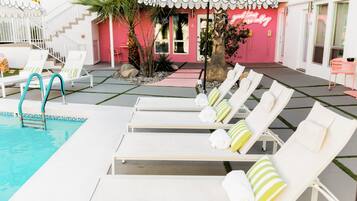 Un área de piscina con sillones de descanso, una escalera de piscina y un edificio de fachada rosa.