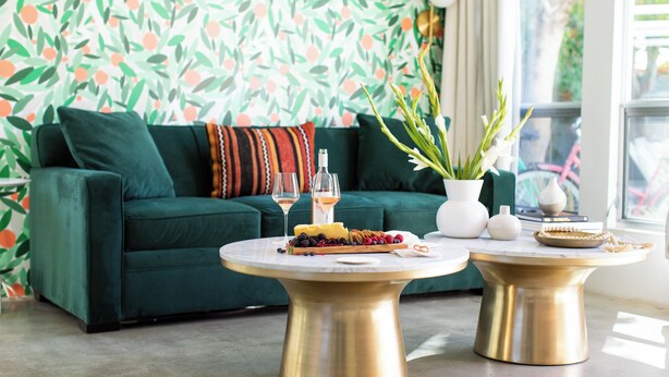 A green sofa with a patterned wallpaper, two gold tables with food and drinks, and a window with a view. - The Wesley by AvantStay (Palm Springs)