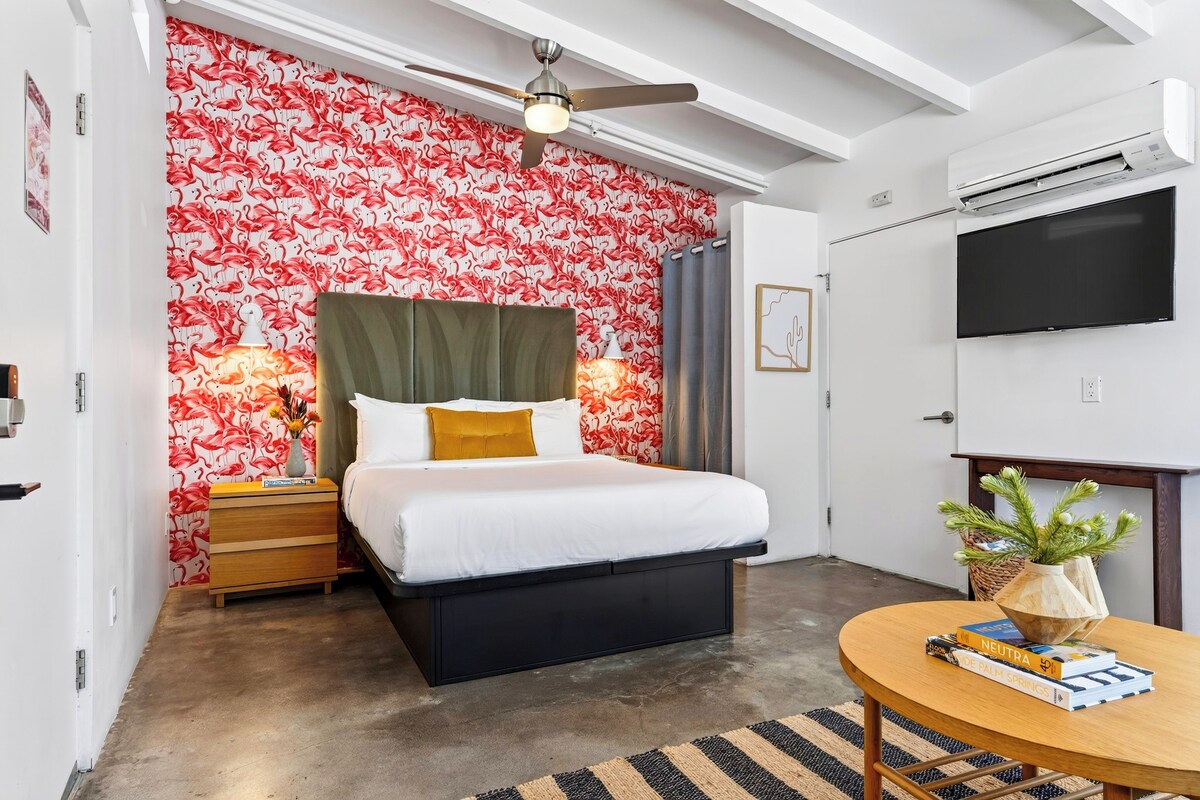 A hotel room with a bed, a bedside table, a lamp, a ceiling fan, a TV, and a small round table with books.