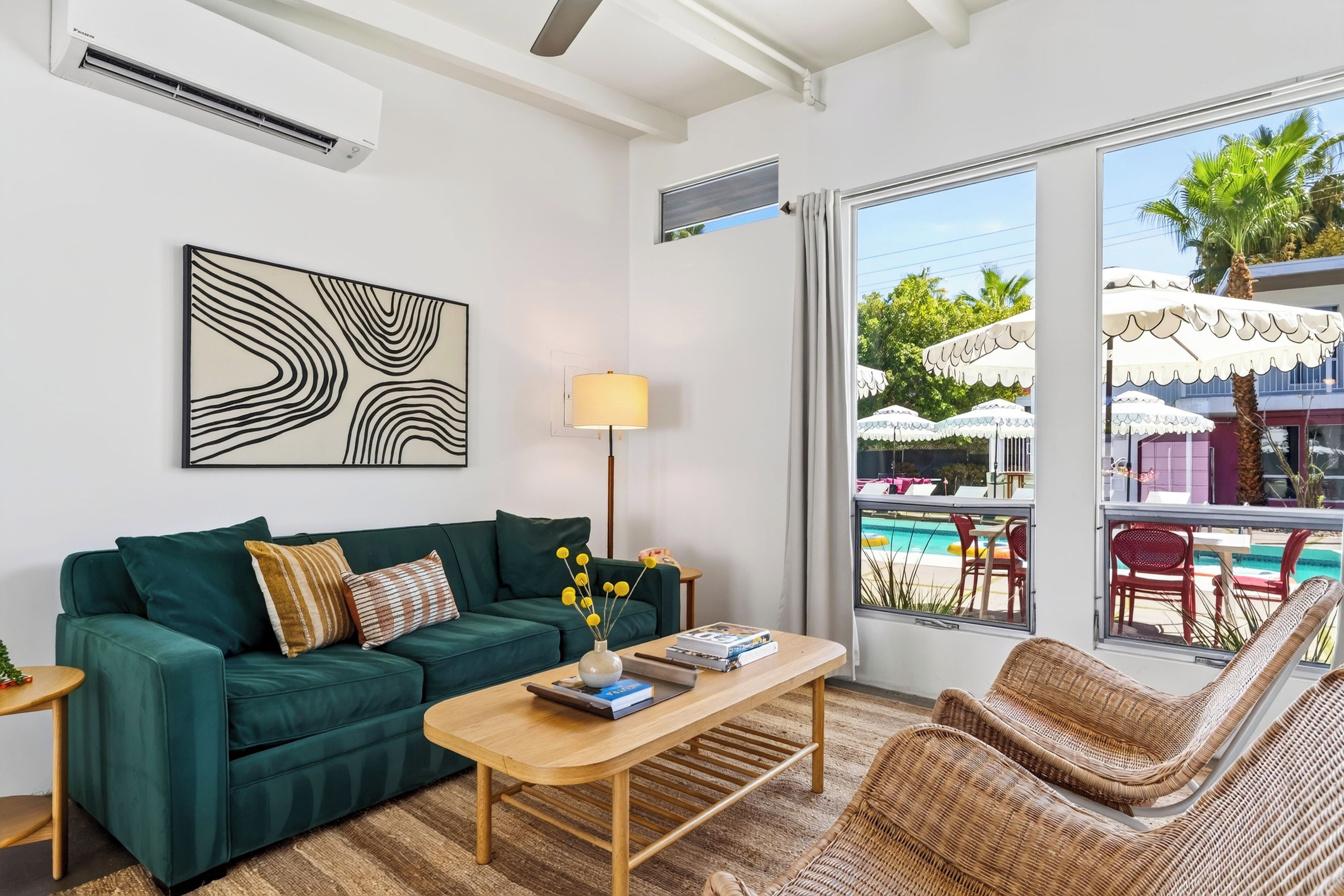 A modern living room with a green sofa, a wooden coffee table, and a wicker chair. A large window offers a view of a pool and outdoor umbrellas.