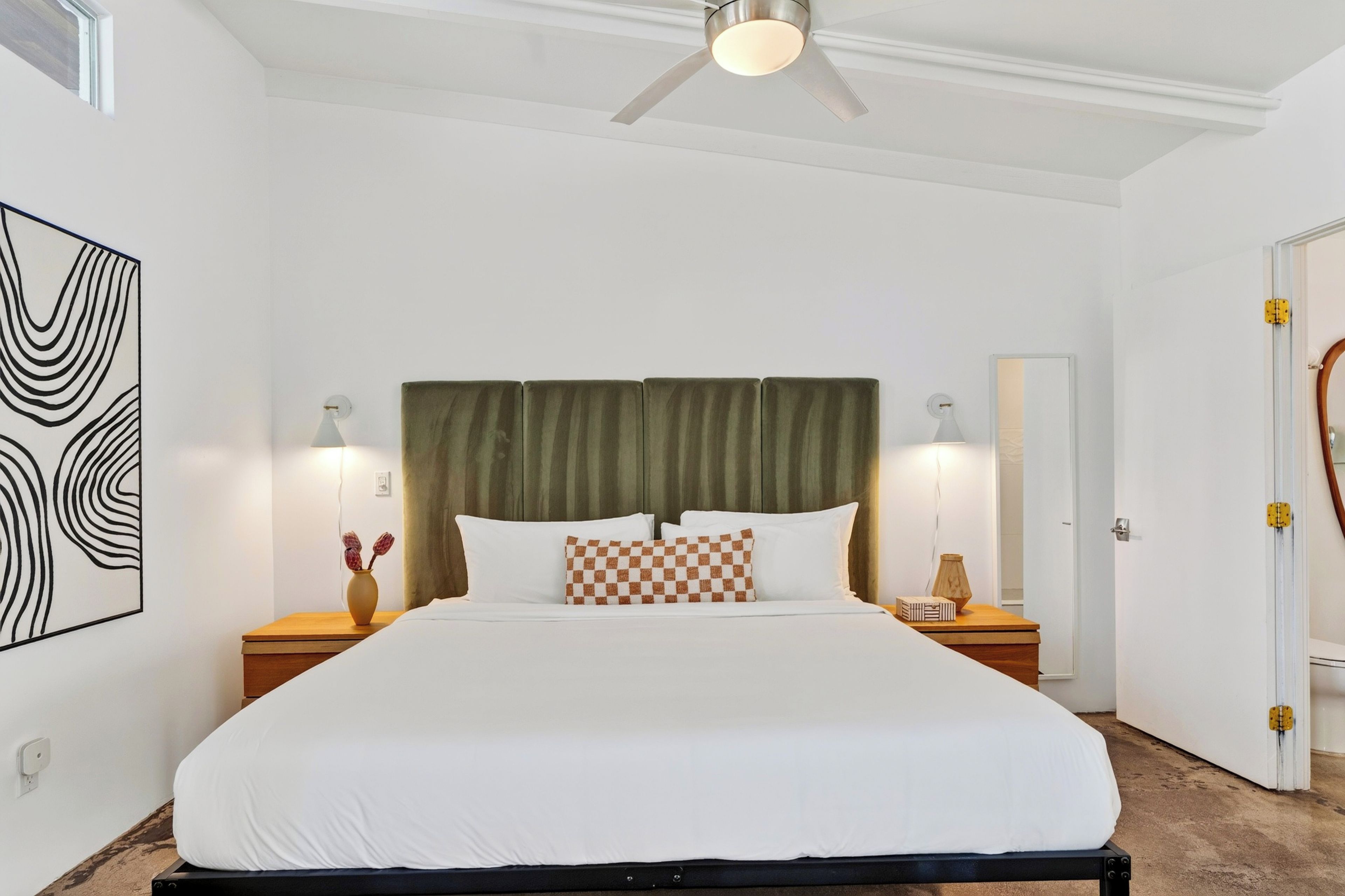 Ein Hotelzimmer mit einem Bett, zwei Nachttischen, einem Deckenventilator, einem großen abstrakten Gemälde und einem Spiegel.