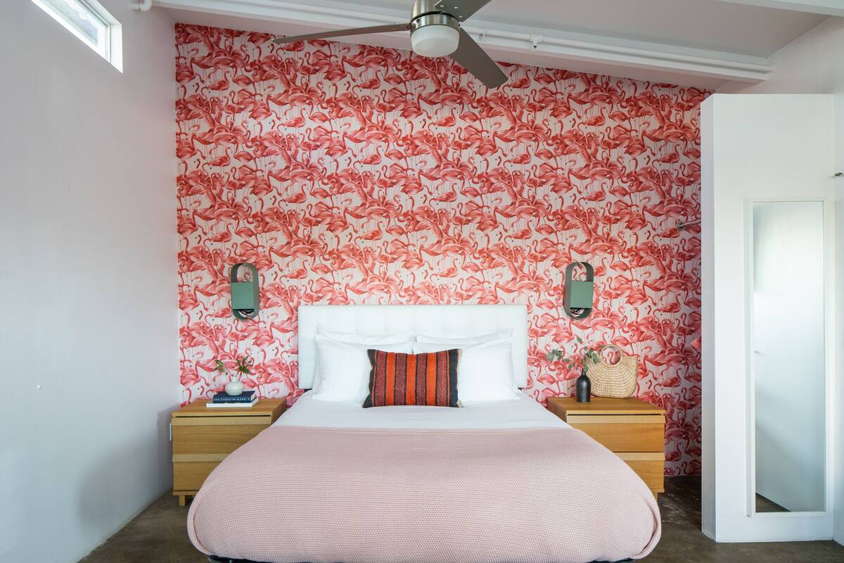 A bedroom with a white headboard, a pink bedspread, and a wall with a flamingo pattern. There are two nightstands with lamps and a small table with a vase.