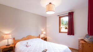 A bedroom with a wooden bed, two bedside lamps, a window with red curtains, and a view of a house outside.
