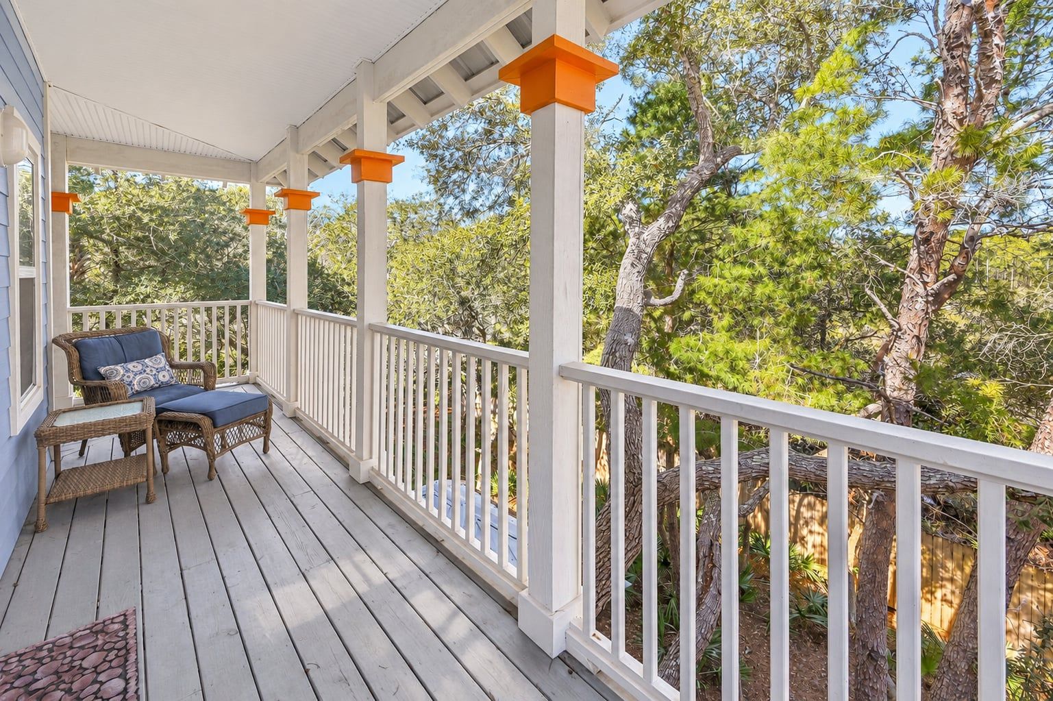 A covered porch with a white railing, a wicker chair with a cushion, and a small table.