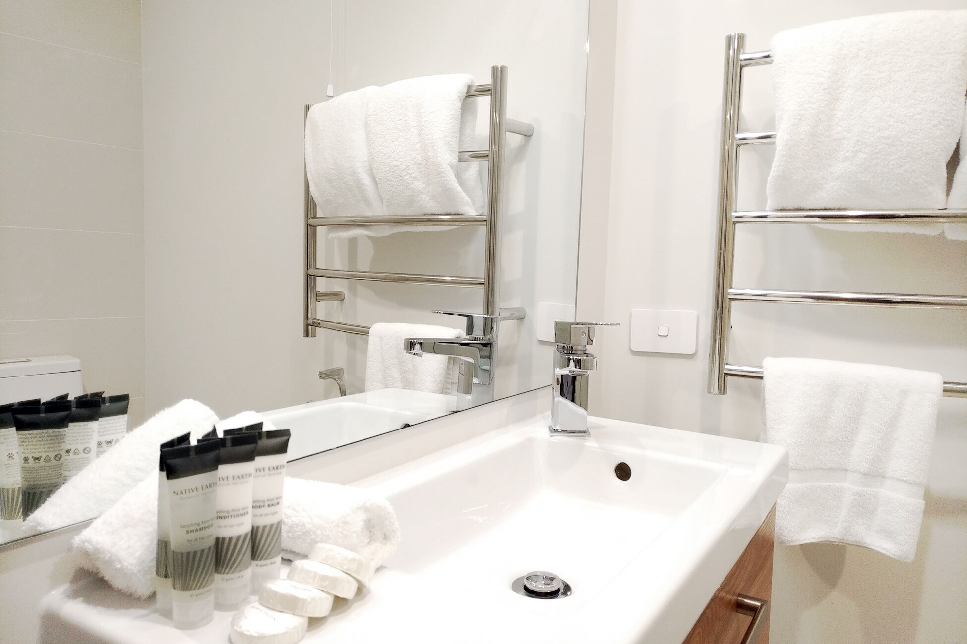 Un baño con un lavamanos blanco, toalleros cromados y un espejo. Hay toallas blancas colgadas en los toalleros y una botella de champú sobre el lavamanos.