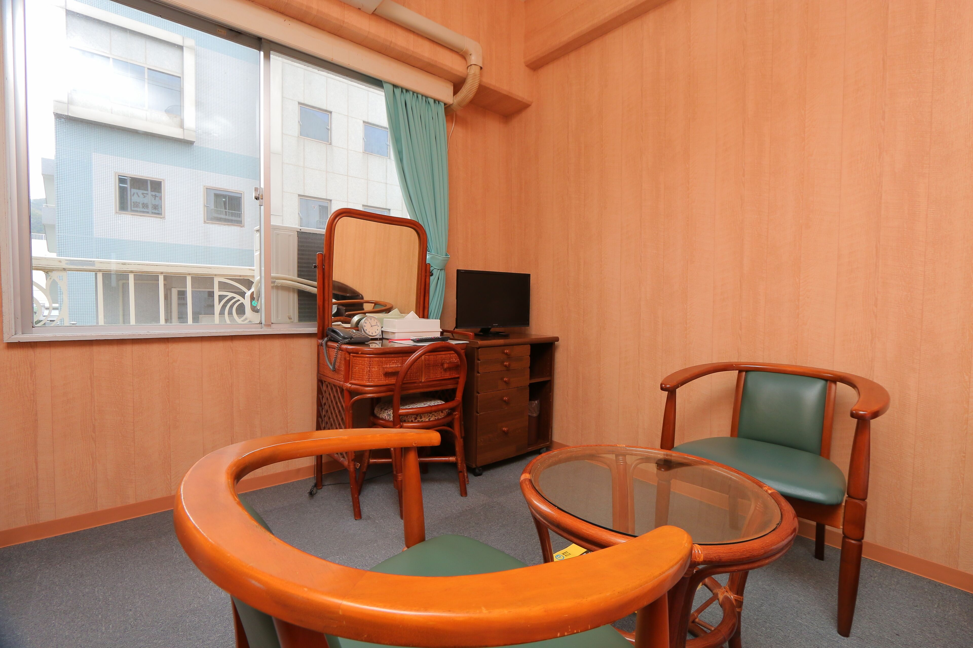 Une chambre d’hôtel avec un bureau en bois, deux chaises, une table en verre, un téléviseur et une fenêtre donnant sur des immeubles.