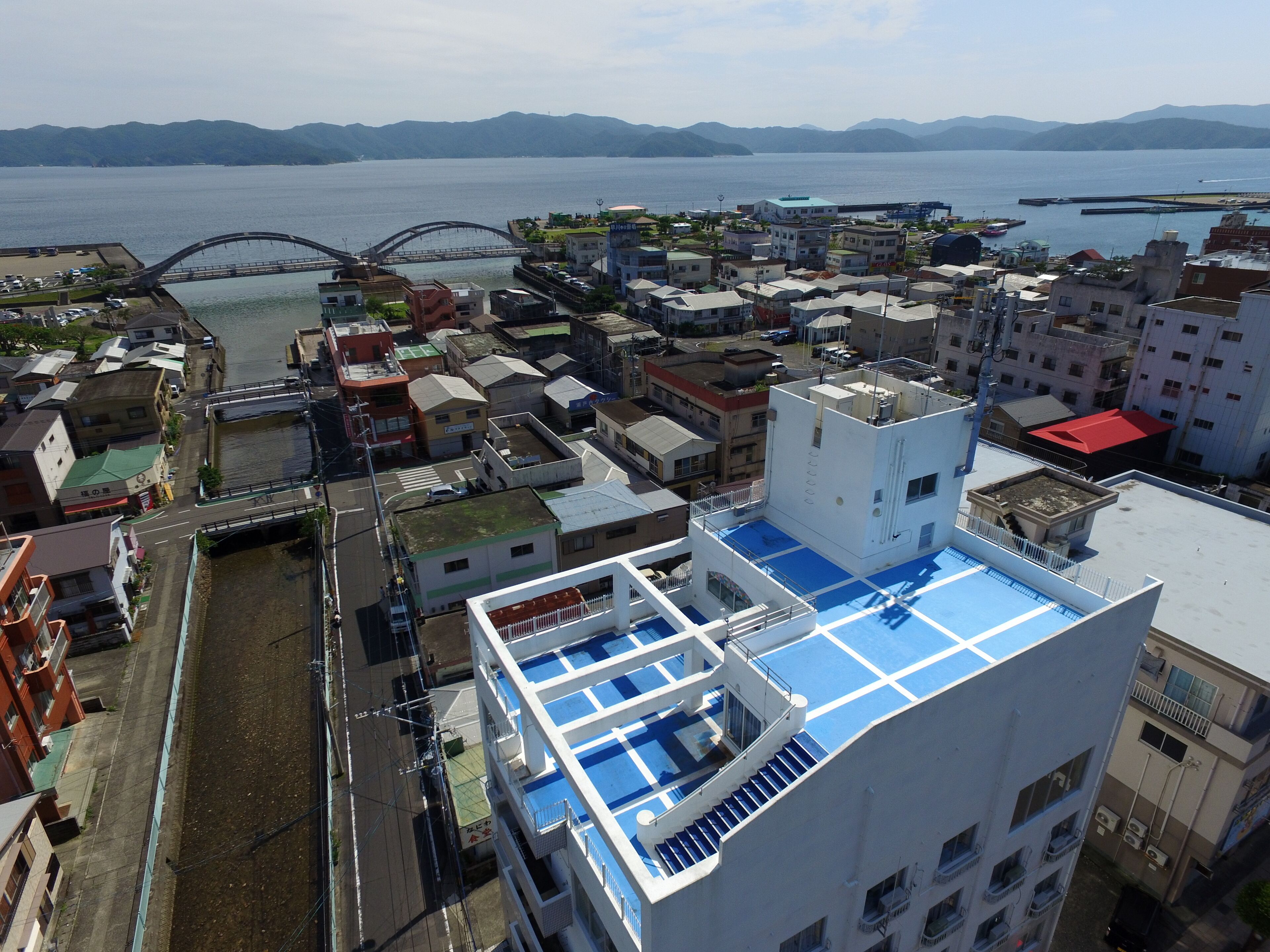 A high-rise building with a blue rooftop terrace, surrounded by other buildings and a bridge over a body of water.
