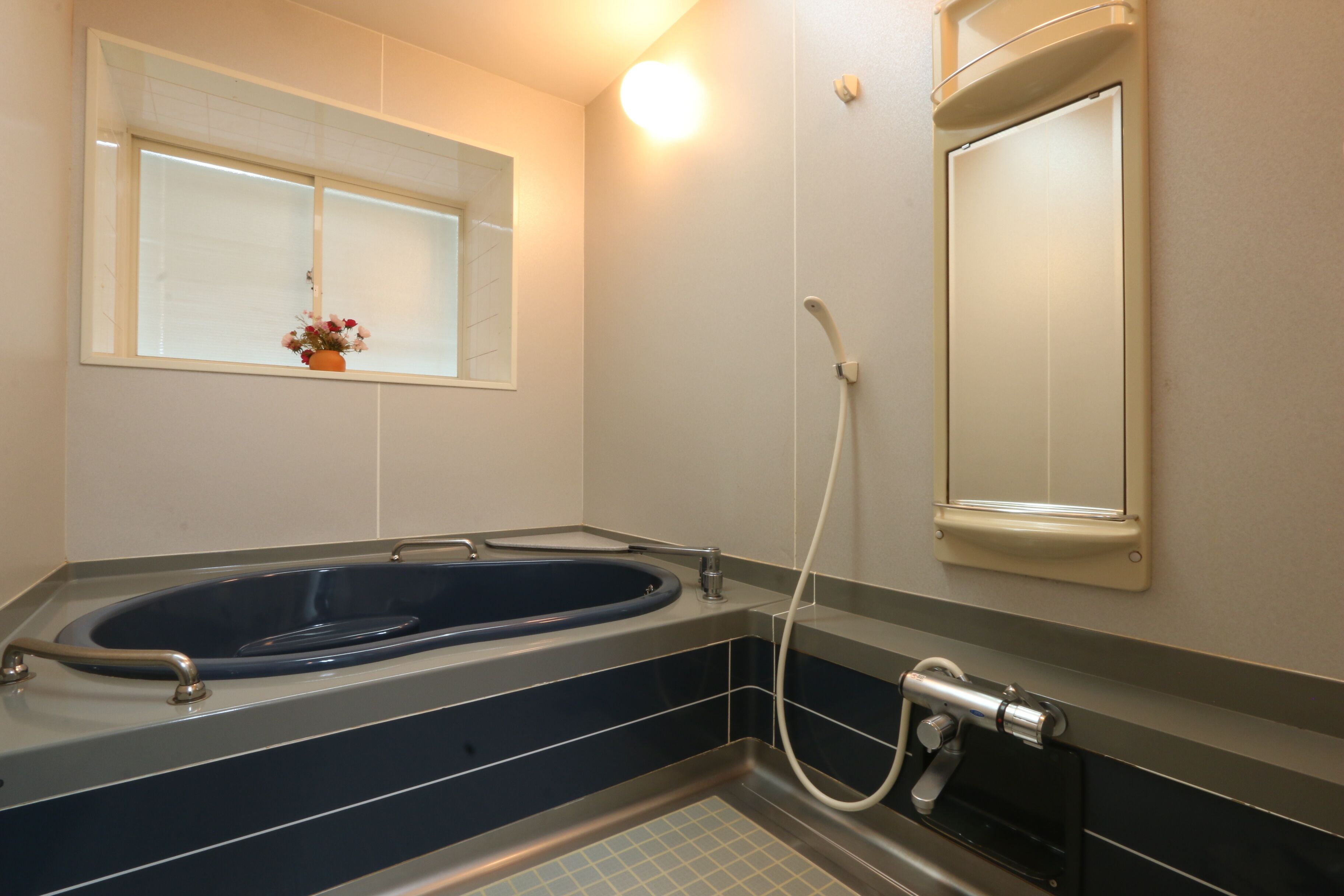 A modern bathroom with a freestanding bathtub, a window with a flower vase, and a wall-mounted mirror.