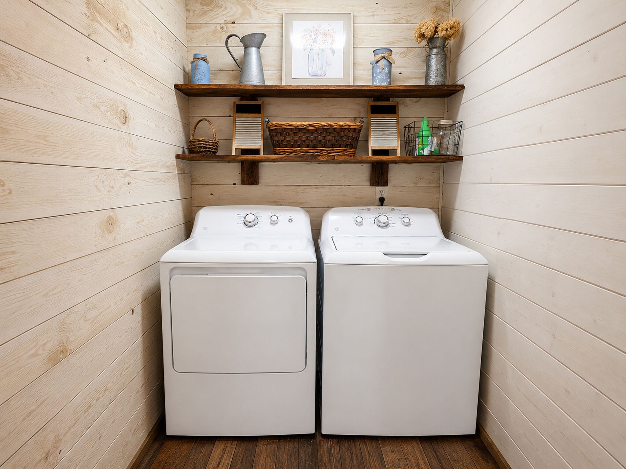 A laundry room with two white front-loading washing machines, wooden walls, and shelves above containing various items.