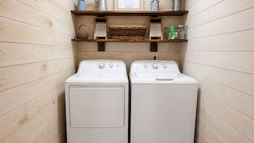 A laundry room with two white front-loading washing machines, wooden walls, and shelves above containing various items.