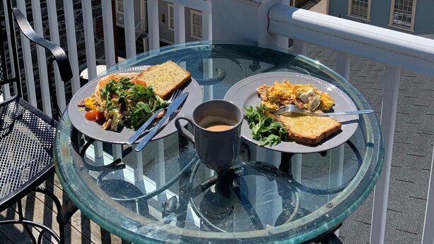 Un balcón con una mesa de vidrio dispuesta para el desayuno, con vista a un puerto con barcos y edificios.