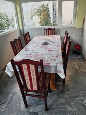 A rectangular dining table with a floral tablecloth, surrounded by six wooden chairs with red and beige upholstery. The room has a tiled floor and large windows with a view of greenery outside. - Apartment in Ždrelac with Balcony, Air condition, WIFI, Washing machine (4834-1) (Ždrelac)