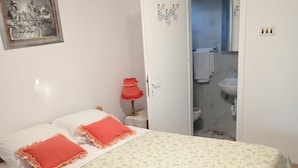 A bedroom with a bed, a nightstand, a red lamp, a door leading to a bathroom, and a wall-mounted picture.