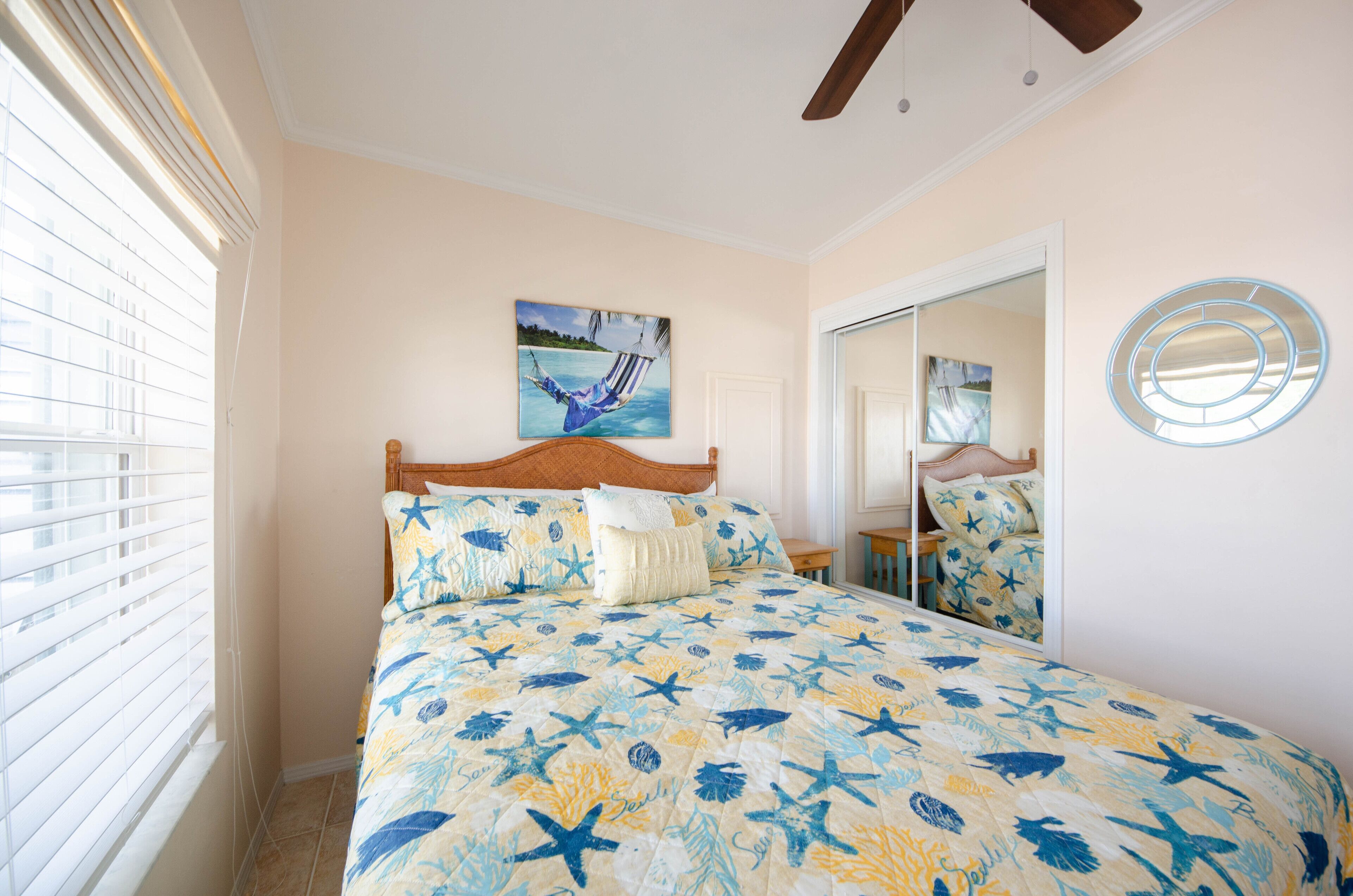 Une chambre à coucher avec un lit, un ventilateur de plafond, une fenêtre avec des stores, un tableau et un miroir.