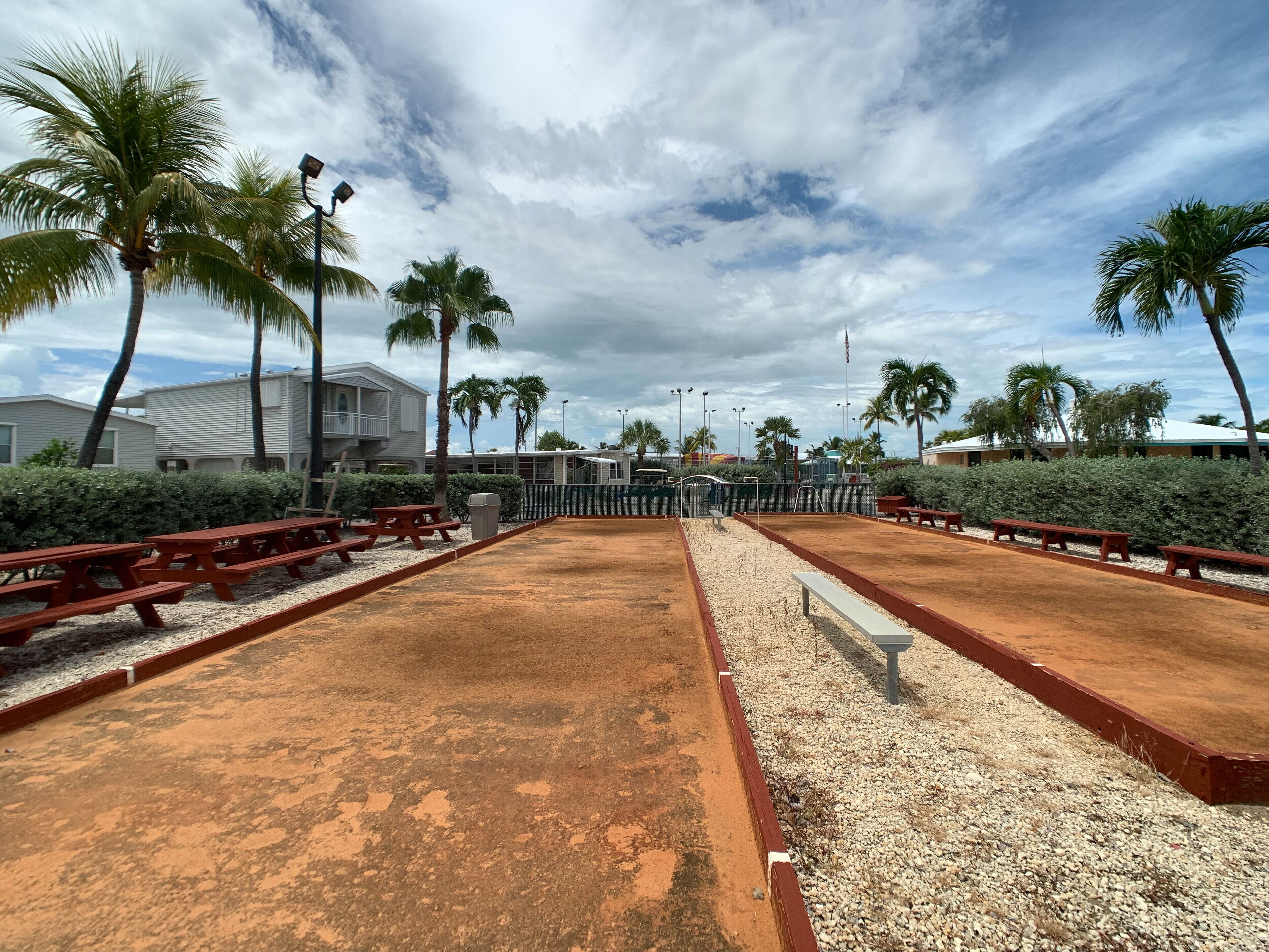 A sandy court with benches and palm trees, surrounded by buildings and a cloudy sky.
