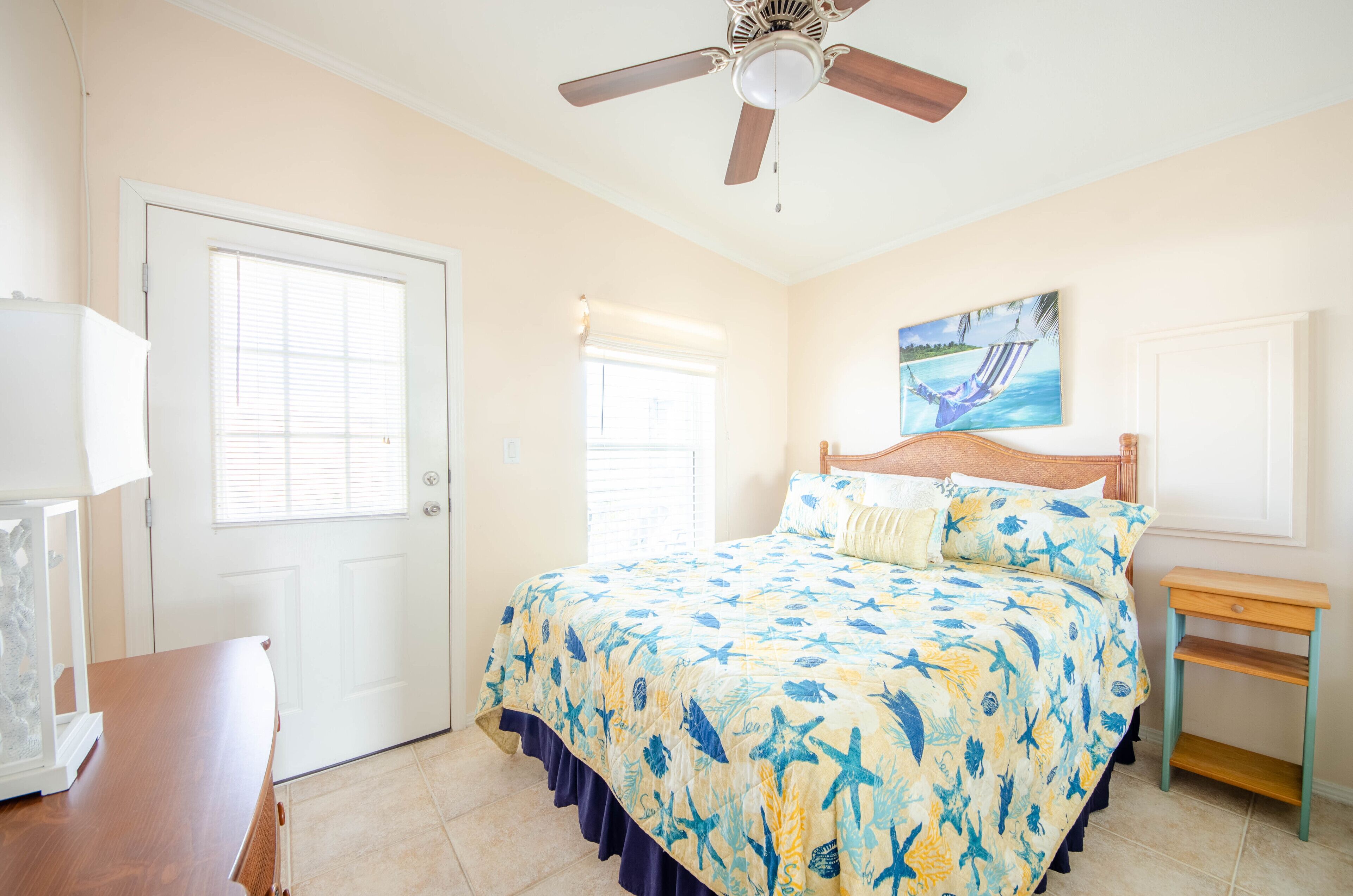 A bedroom with a bed, a ceiling fan, a window, and a painting on the wall.