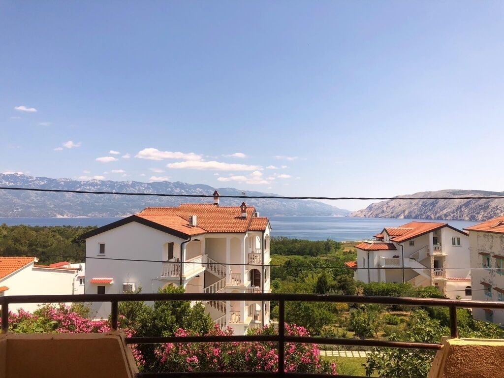 Un área residencial costera con edificios blancos y techos de tejas rojas, cielo despejado y vistas al mar y montañas a lo lejos.