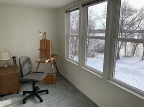 Une pièce avec un bureau, une chaise et un chevalet, de grandes fenêtres donnant sur un paysage enneigé.