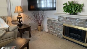 A living room with a flat-screen TV mounted on the wall, a stone fireplace, a sofa, a coffee table, and a lamp. - ARTISTS & ANGLERS INN - game room - home of Madison Lake Paddlefish!! (Madison Lake)