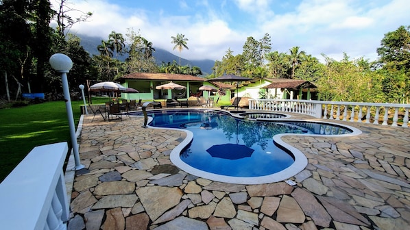 Una piscina con borde de baldosas de piedra, rodeada de sillones de descanso y sombrillas, con vista a una montaña y abundante vegetación al fondo.