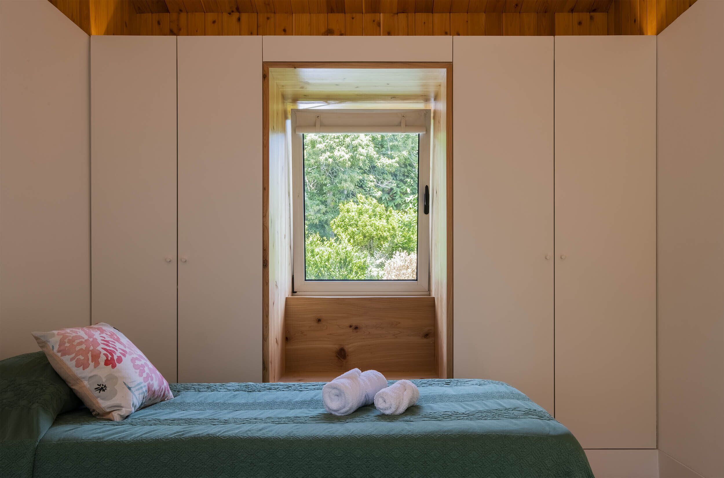 Ein Schlafzimmer mit einem Bett, einem Kopfkissen und gerollten Handtüchern. Ein Fenster bietet Blick auf grüne Natur.