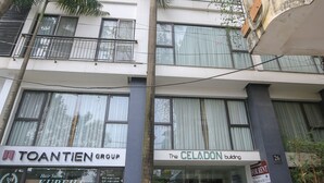 Un immeuble à plusieurs étages abrite un salon de coiffure appelé « TOANTIEN GROUP » et « KUREHA » au rez-de-chaussée. On peut y voir des plantes en pot et un scooter garé à l’extérieur.