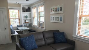 A living room with a brown leather sofa, two blue pillows, a coffee table with a remote control, and a dining area with a white table and chairs.