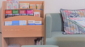 A wooden bookshelf with travel guides and books, a grey sofa with a patterned pillow, and a plastic container on the floor. - SC1 Kaikanoyado (Osaka)