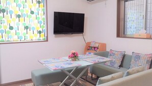 A cozy living room with a sofa, a small table with a vase of flowers, a flat-screen TV mounted on the wall, and a window with a view of the outside. - SC1 Kaikanoyado (Osaka)