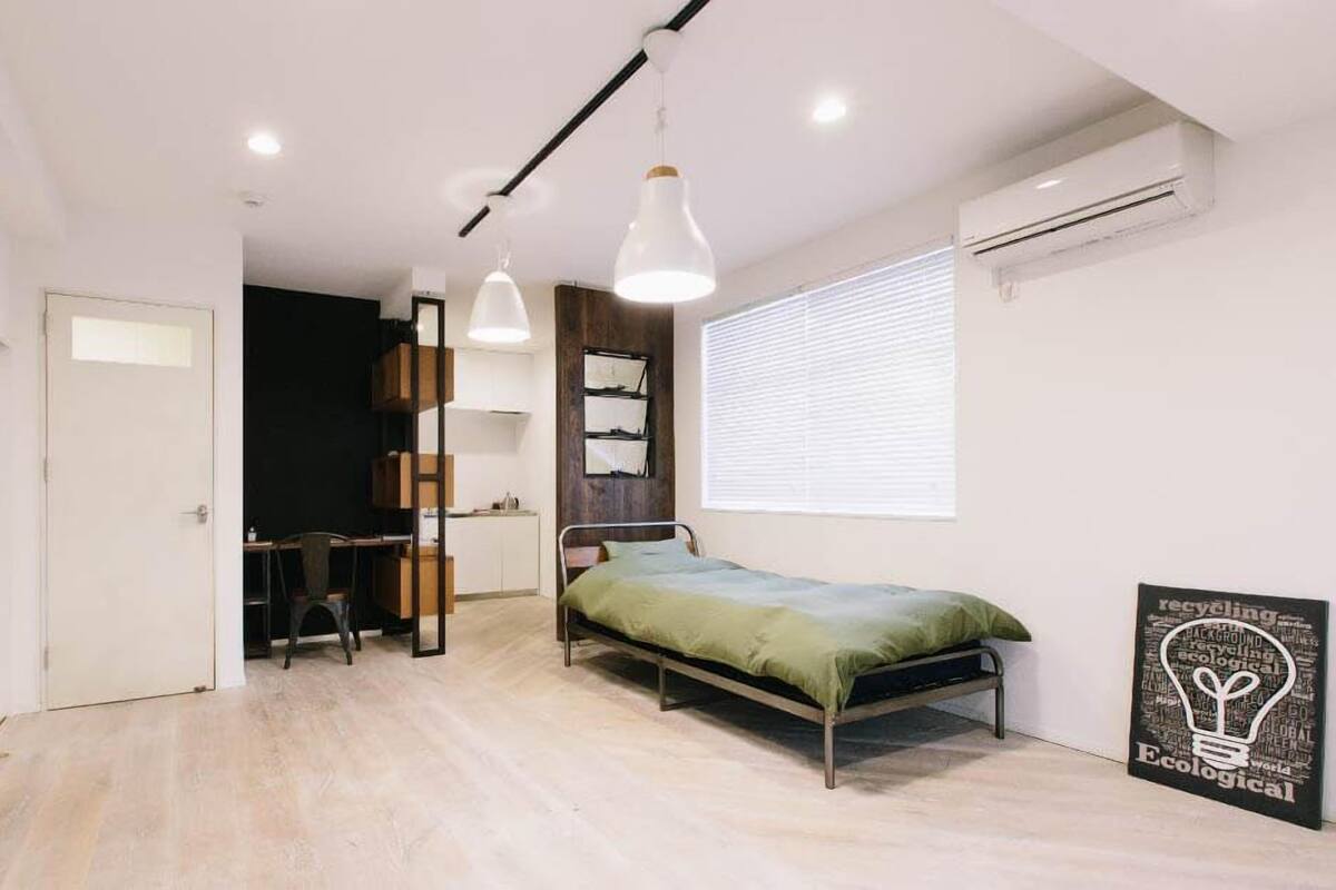 A compact bedroom with a bed, a desk, and a chair. There is a window with blinds, a wall-mounted air conditioner, and a framed poster on the wall.