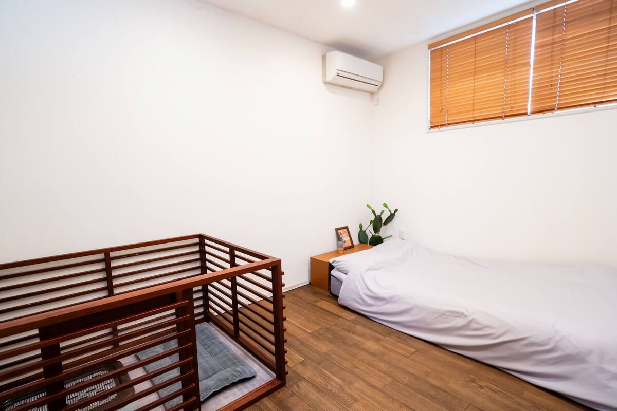A bedroom with a wooden crib, a bed, a nightstand with a cactus, and a window with blinds.