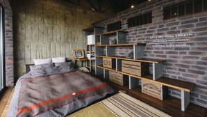 A bedroom with a brick wall, a bed with a gray and orange striped blanket, wooden shelves, and a sign on the wall. - Coton (Kanonji)
