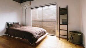A bedroom with a bed, a window with blinds, a ladder, and a trash can.