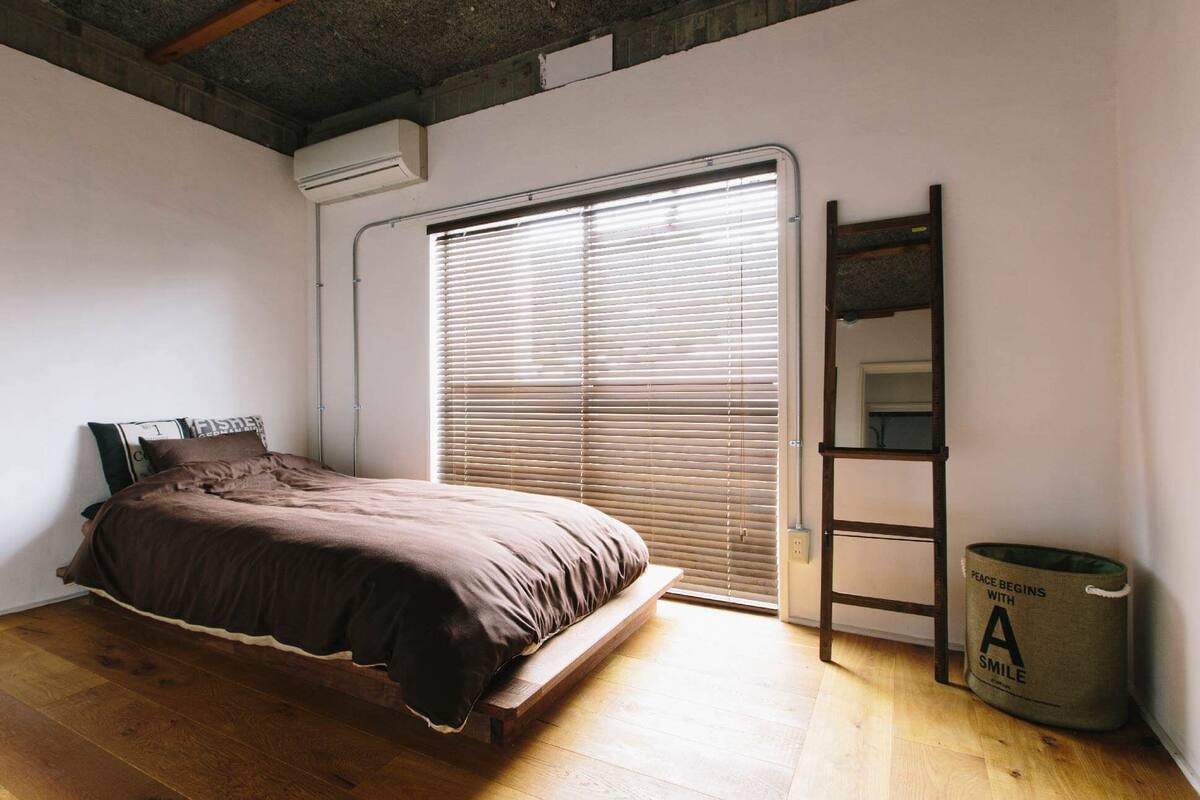 A bedroom with a bed, a window with blinds, a ladder, and a trash can.