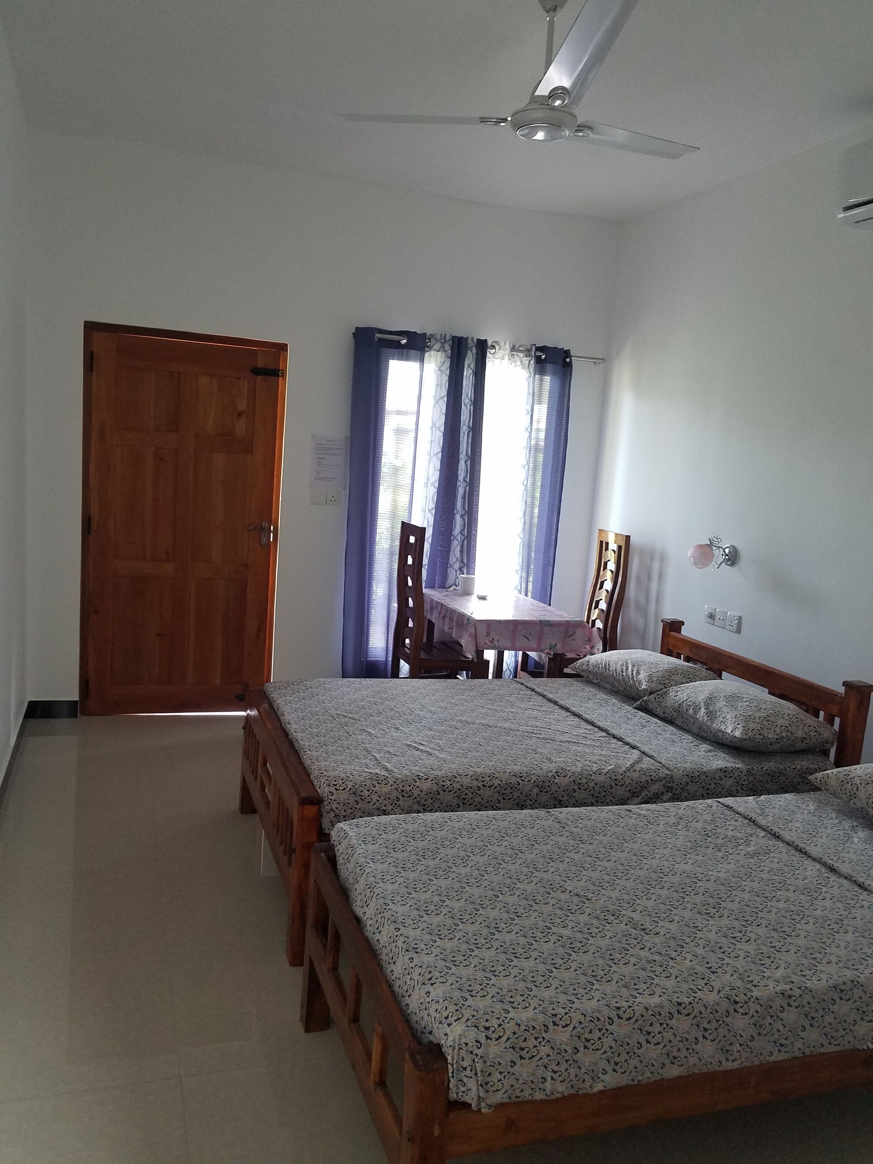 A room with two wooden beds, a ceiling fan, a wooden door, and a table with chairs.